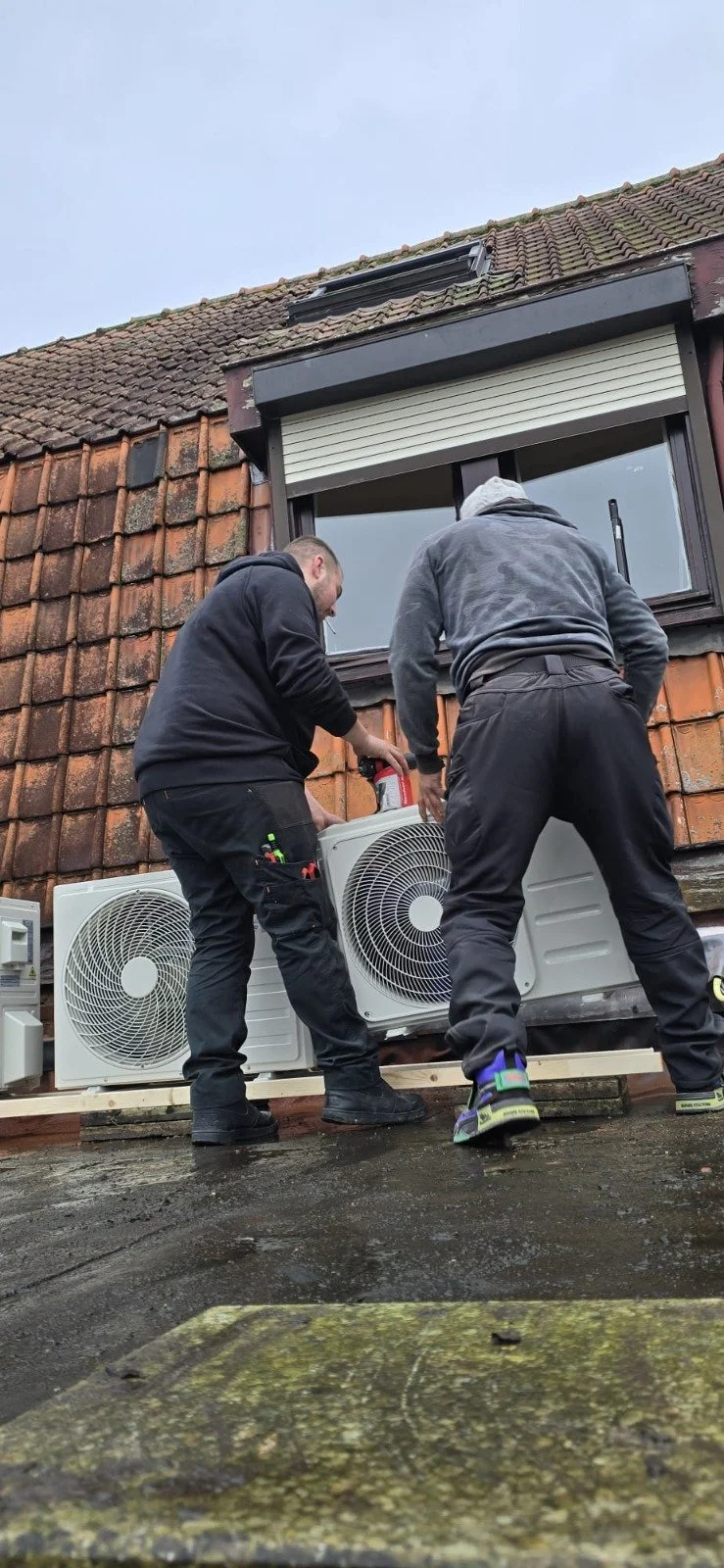 Twee monteurs installeren een buitenunit van een airconditioningsysteem op een dak onder een raam met een rolluik.