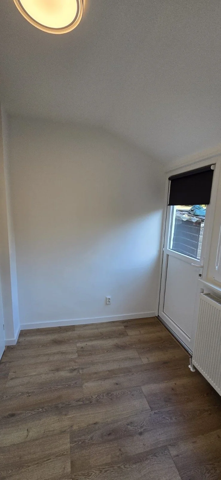 Leeg kamertje met witte muren, een raam met een zwarte rolgordijn, houten vloer, en een radiator onder het raam.