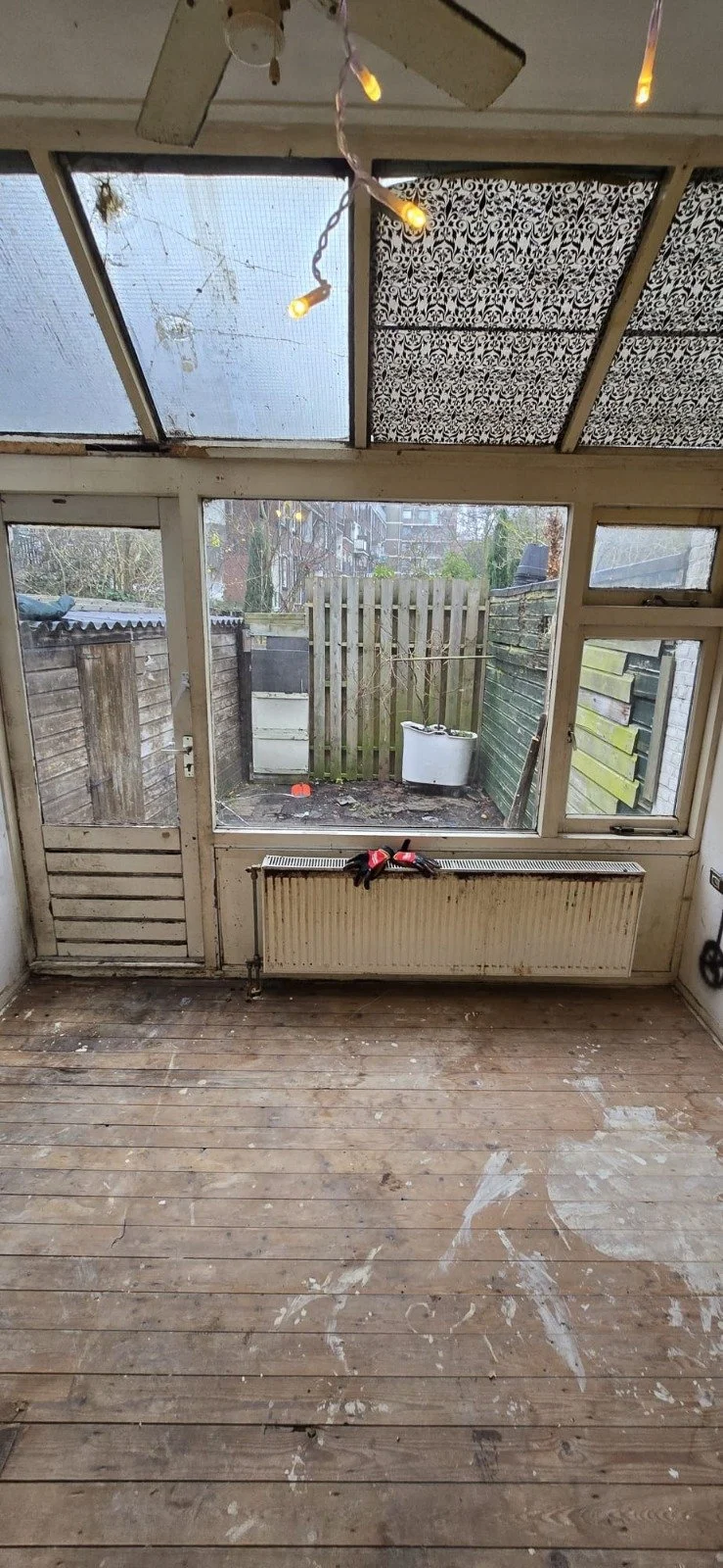 Binnenruimte met houten vloer, kapotte plafond met losse plafondfan en stringer met lichten, grote ramen en deur naar tuin met houten schutting en vuil, radiateur met werkhandschoenen.