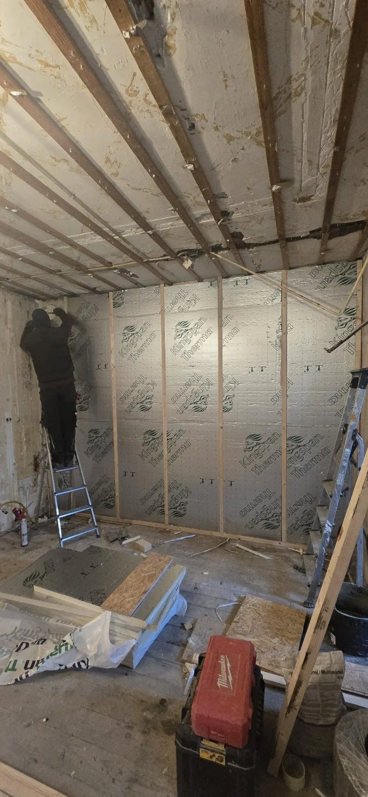 Binnenkant van een kamer onder renovatie, met iemand die zich bezighoudt met de verf- of gipswerk aan de muur, met geïsoleerde wanden en enkele ladders en bouwmaterialen op de vloer.