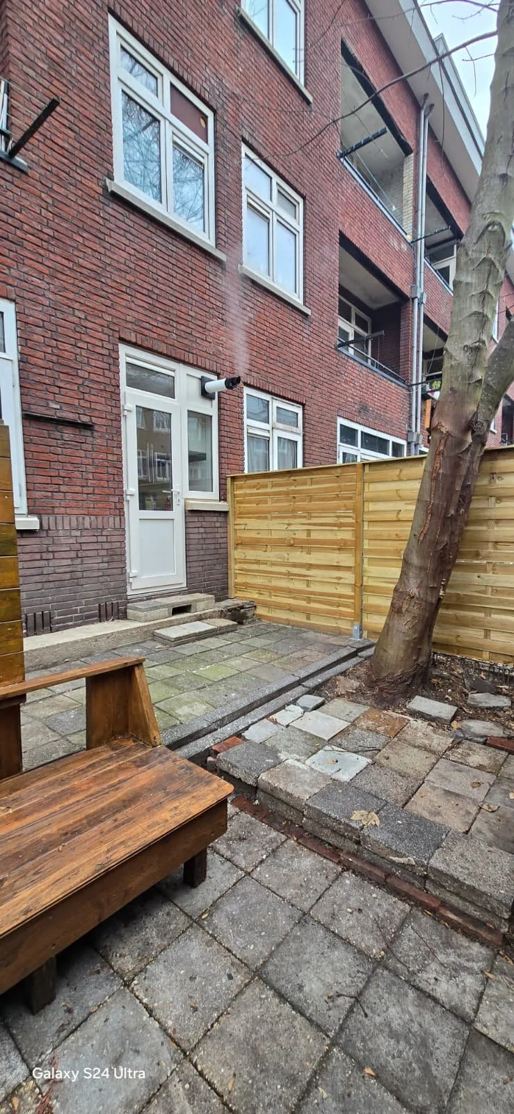 Een kleine binnenplaats met een houten bank, een boom, een houten schutting, een stenen vloer en een gevel van een rood bakstenen gebouw met meerdere ramen.