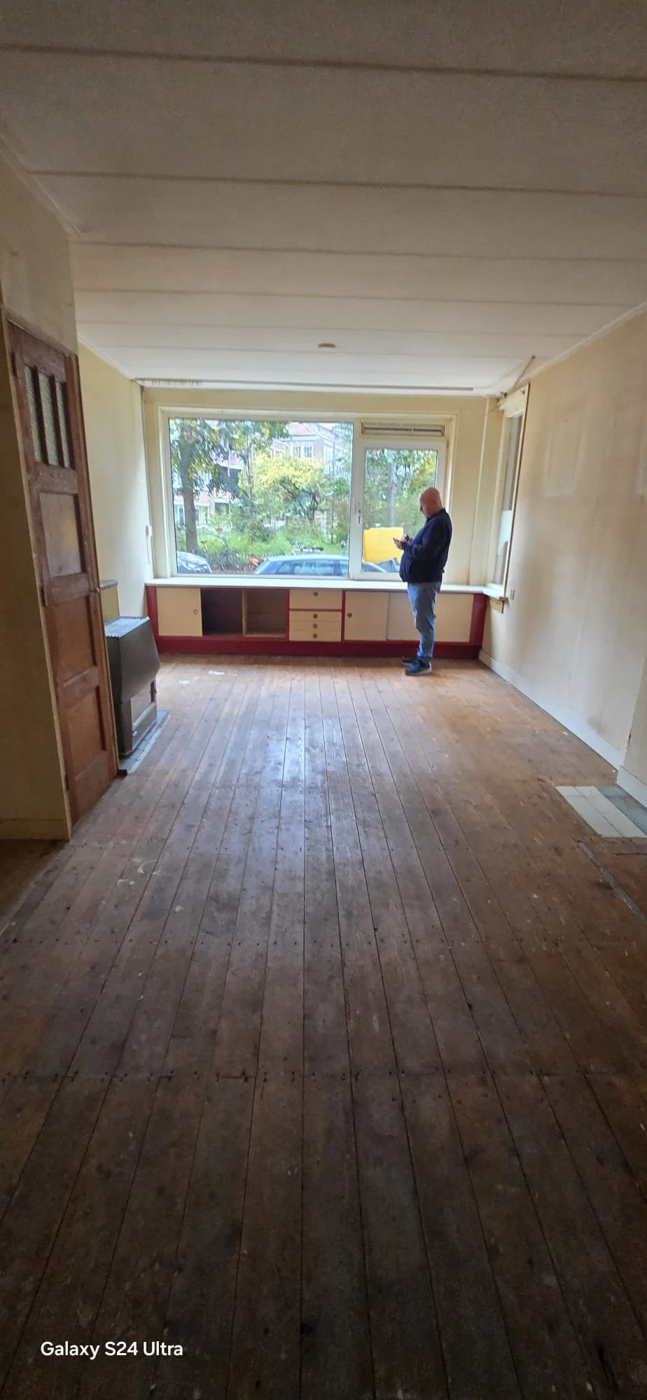 Lege woonkamer met houten vloer, grote ramen, en man die op zijn telefoon staat.