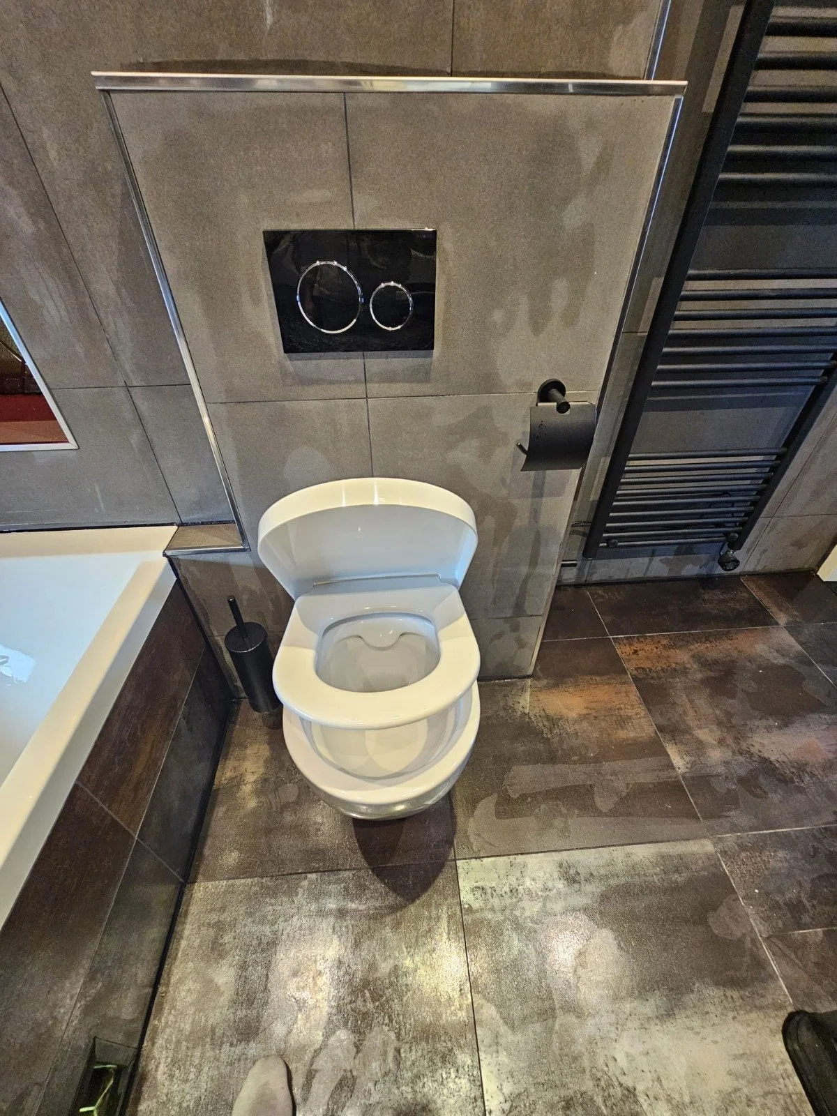 Wandtoilet met zwart bedieningspaneel en toiletborstel in een badkamer met donkere tegels.