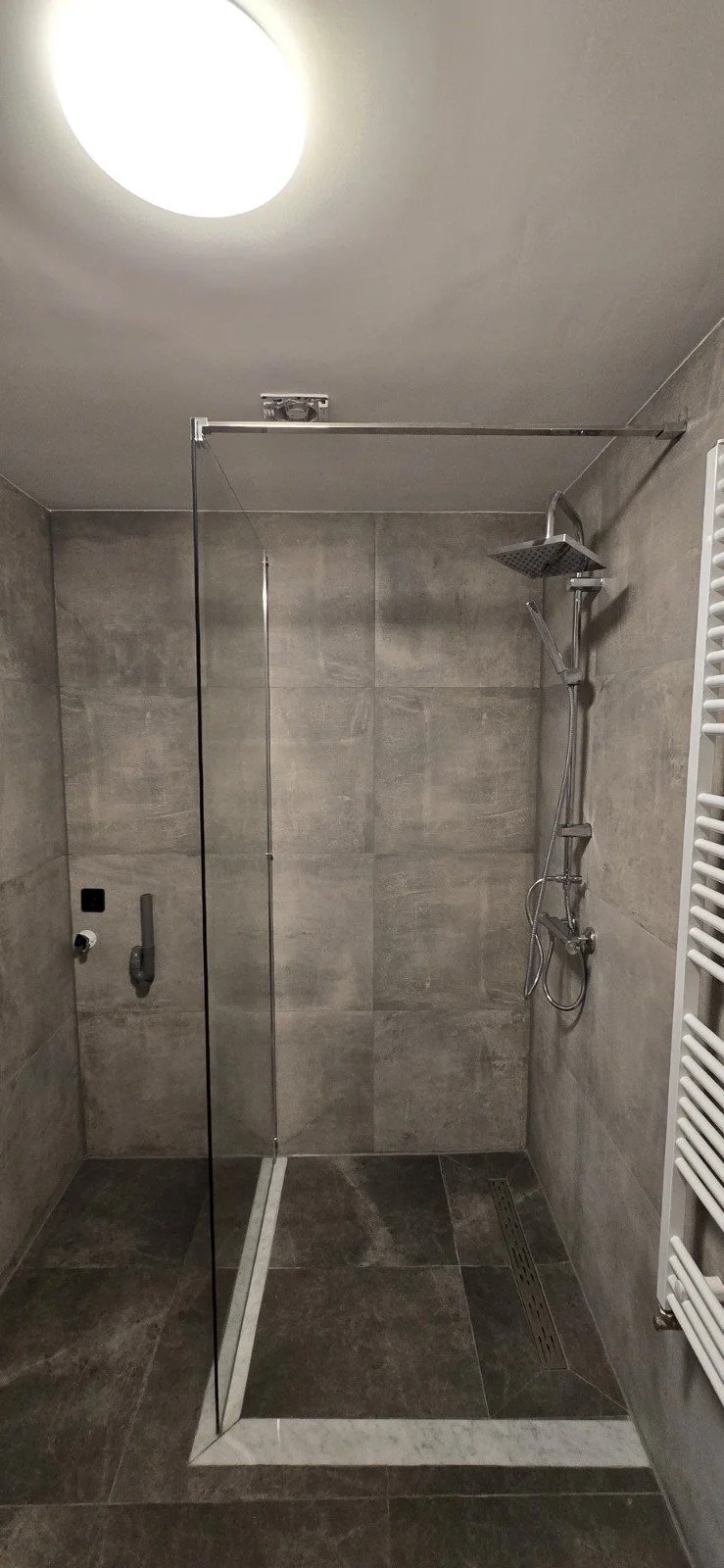 Inloopdouche met grijze keramische tegels, regendouche, handdouche, glazen schuifwand, centrale afvoer, geplafondverlichting, witte radiator