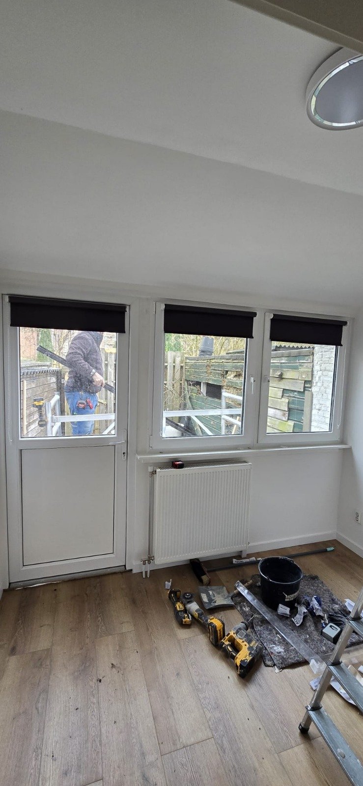 Binnenruimtes met grote ramen, een deur, een radiatoren en gereedschap zoals een boormachine en een emmer, terwijl iemand buiten bouwt of klust.