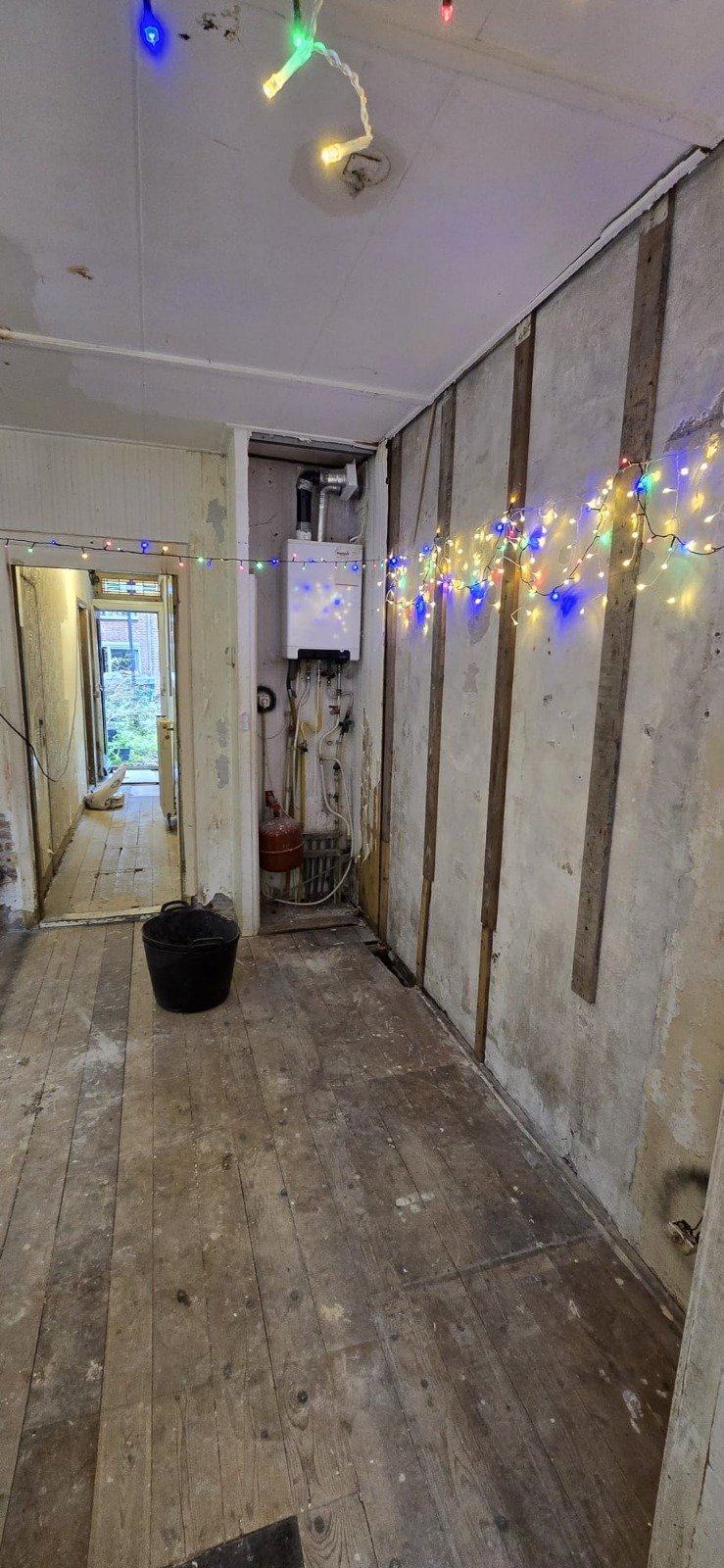 Een kamer in aanbouw met ongeschuurde houten vloer, decoratie met gekleurde kerstlichtjes aan de muur en plafond, en een waterboiler op de muur.