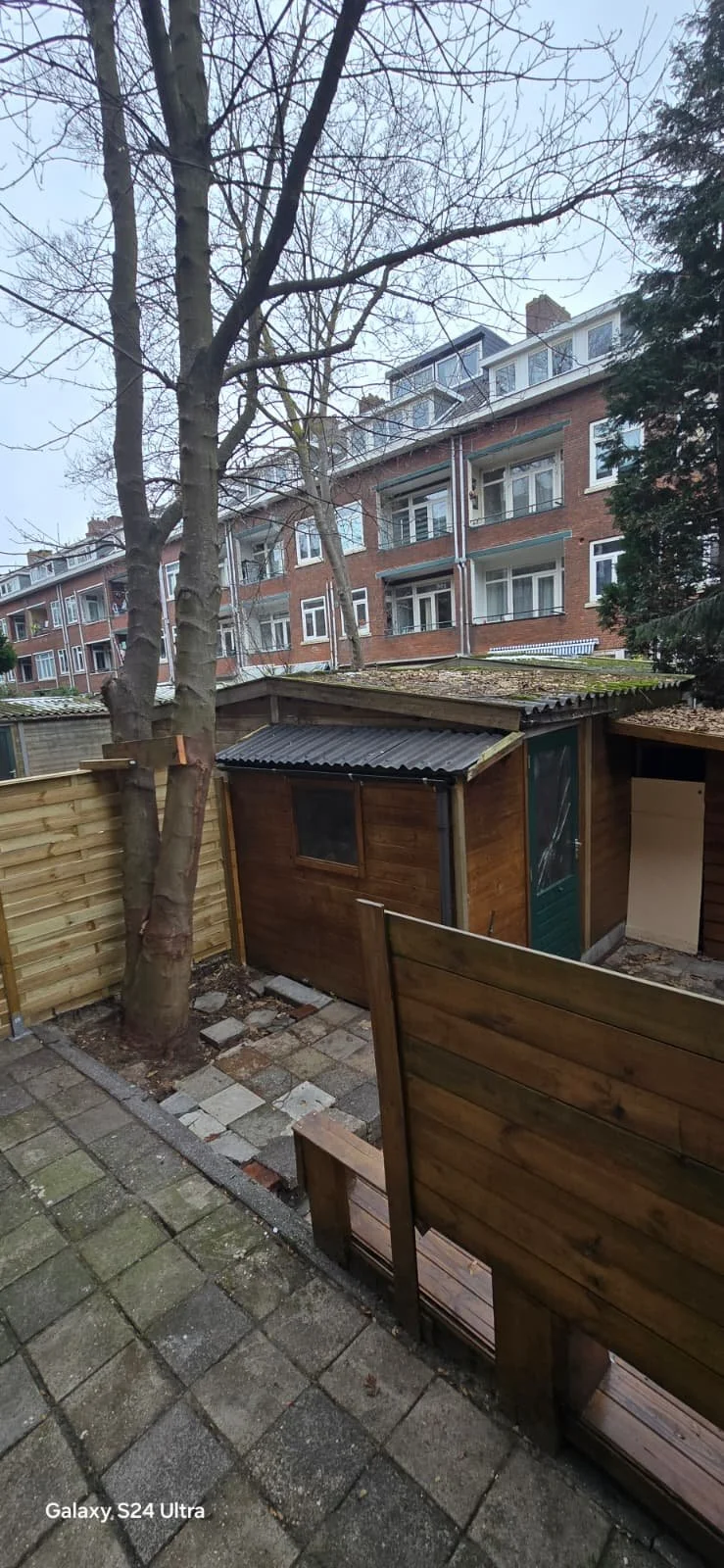 Een zomertuin met een kleine houten schuur, omgeven door een houten hek, met een stenen tegelpad en een boom met kale takken. Achter de tuin is een appartementsgebouw met meerdere verdiepingen.