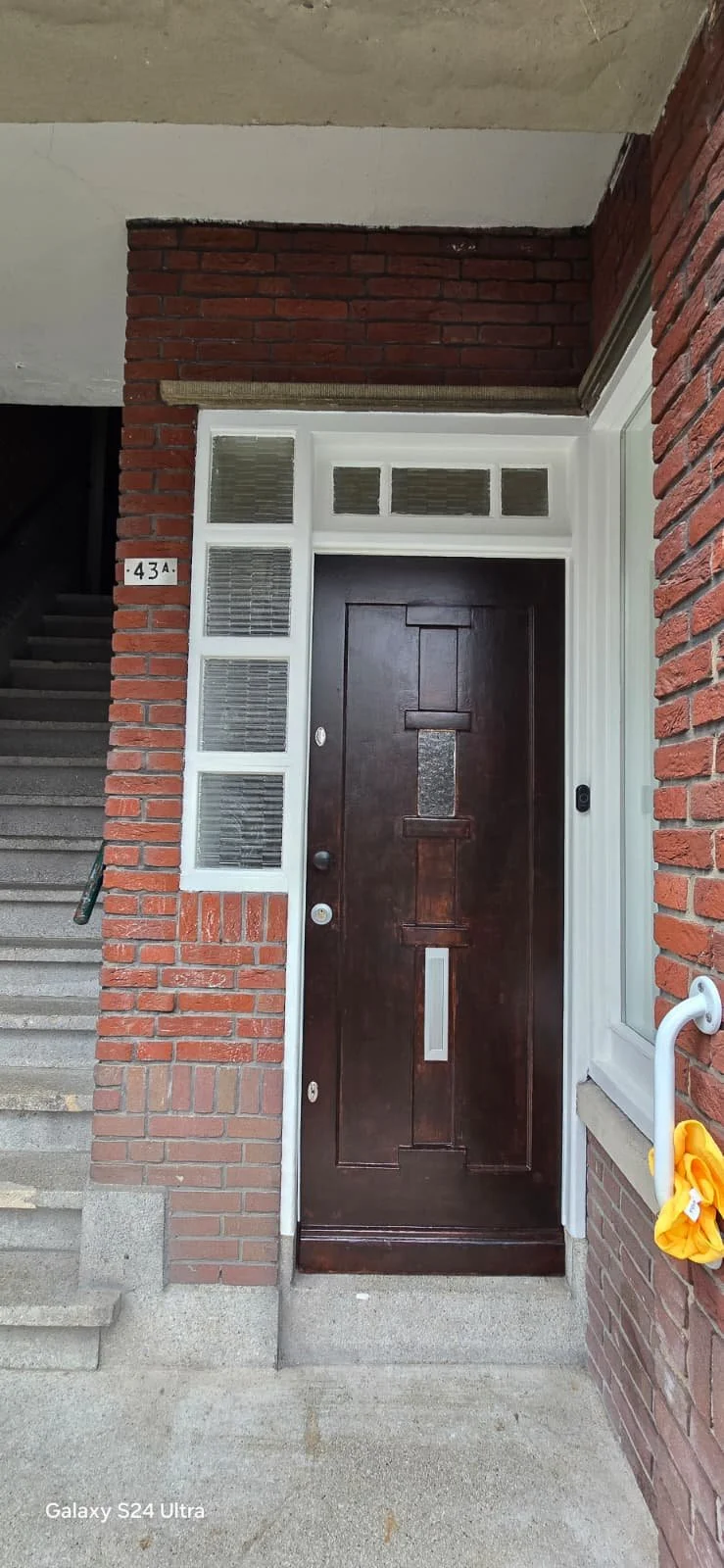 De ingang van een appartement met een houten deur, omringd door een witte lijst en glazen blokken, met een stenen trap en een huisnummer 43A zichtbaar.