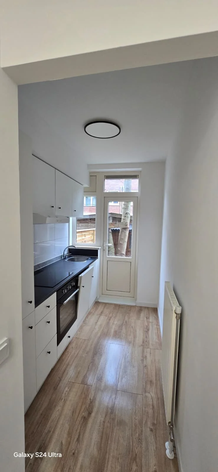 Een kleine keuken met witte kasten, een zwart aanrechtblad, een oven, een wastafel en een deur naar een tuin, met houten vloer en een plafondlamp.