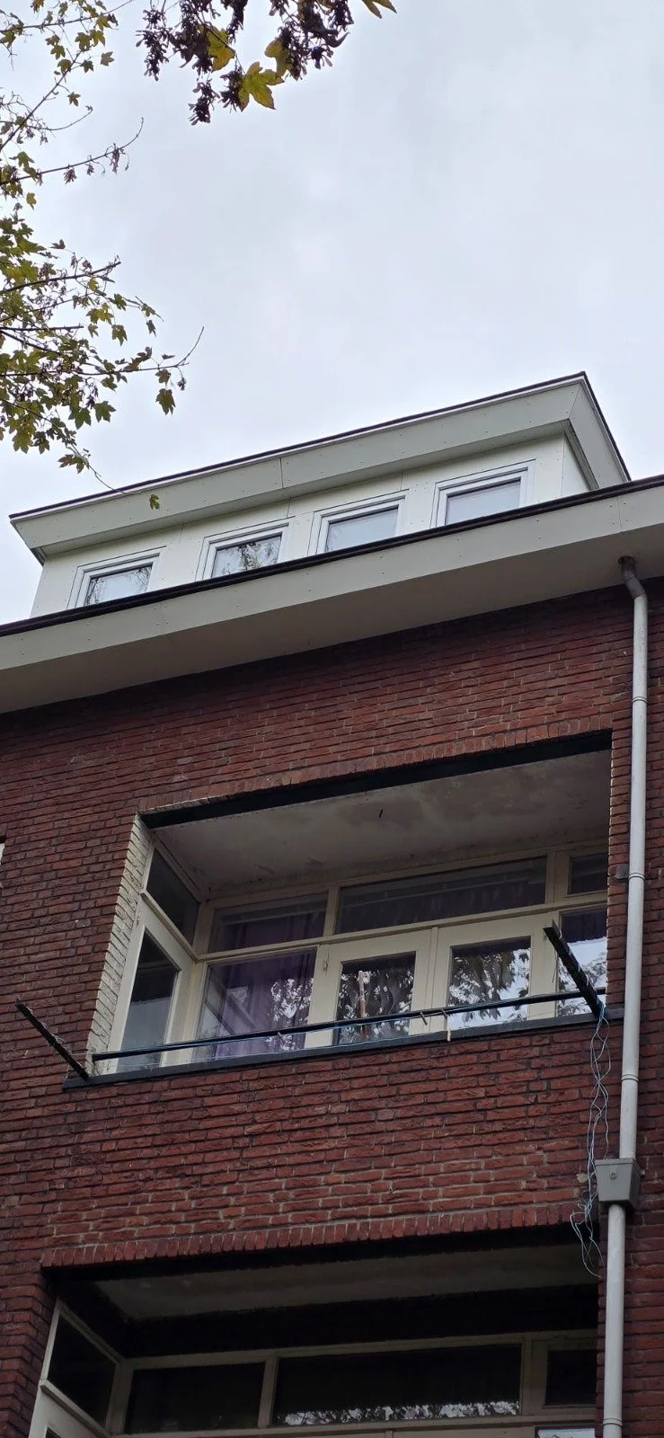 Een woongebouw met een gevel van rode baksteen en houten ramen, bovenin een witte aanbouw met drie ramen en een blauwe hemel erachter.