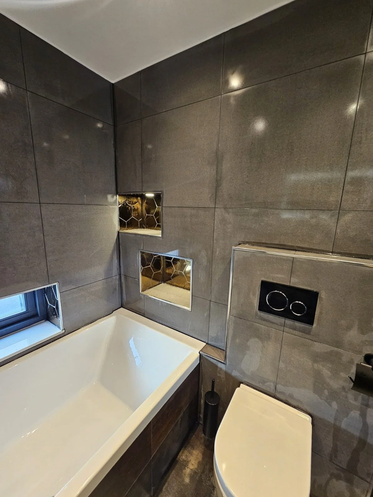 Badkamer met donkergrijze betegelde muren, witte badkuip, een toilet, een zwart toiletrolhouder en een zwart toiletborstel. Er zijn drie vierkante spiegels ingebouwd in de muur.