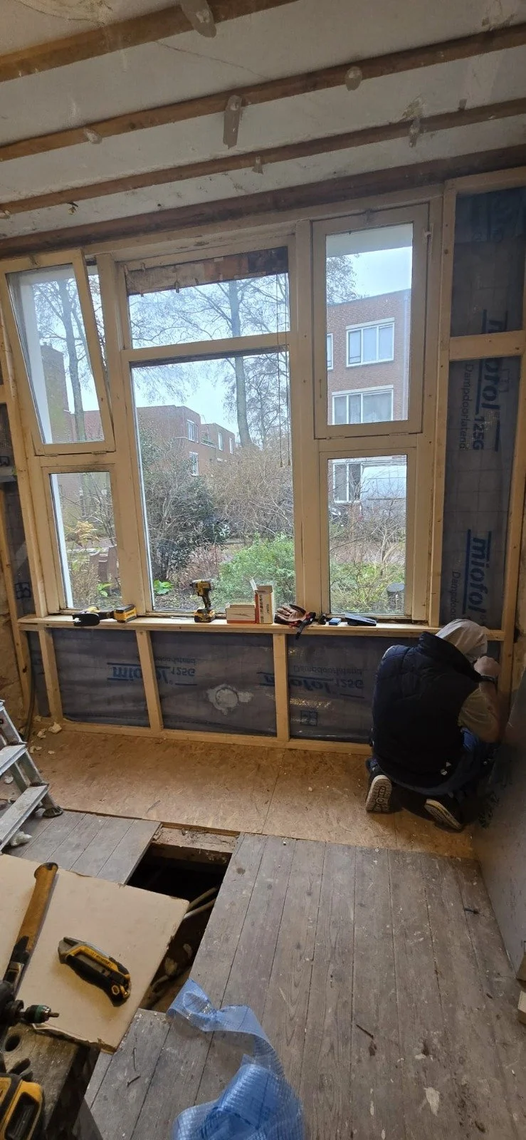 Binnenzijde van een kamper met een grote raamopening, iemand die aan het werk is, bouwmaterialen en gereedschap op de vloer.