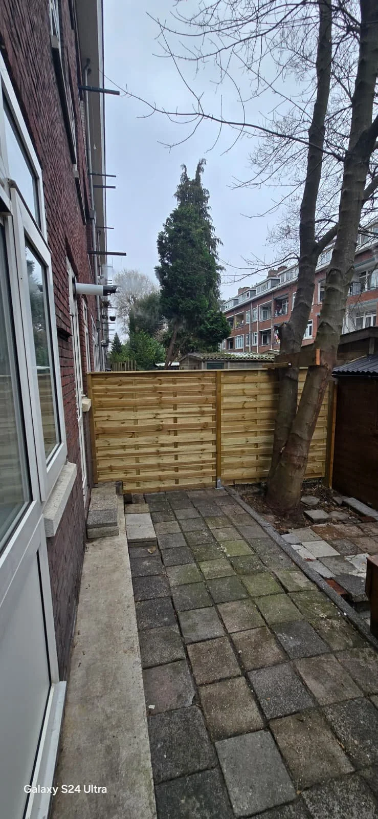 Een tuin met een houten schutting, een boom en gevels van appartementen op de achtergrond.