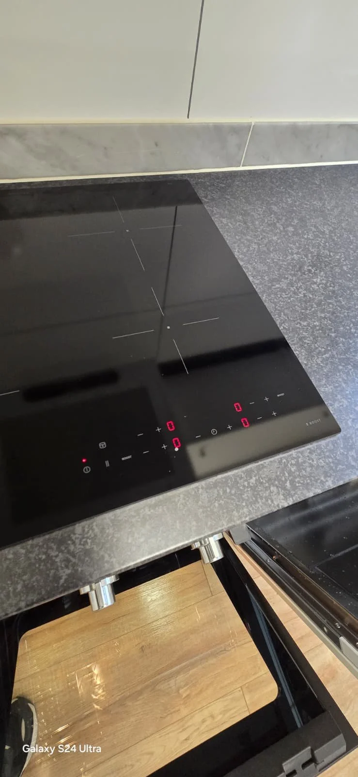 Een zwart keramisch kookplaat met digitale bediening en touch controls, geplaatst op een grijze keukentegel en houten vloer.