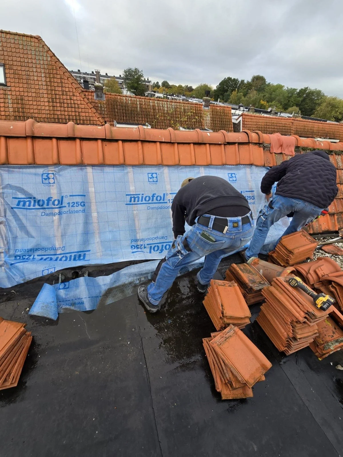 Twee klusploegen werken samen op een oud schuindak, met stapels nieuwe terracotta dakpannen en bouwmaterialen rondom hen.