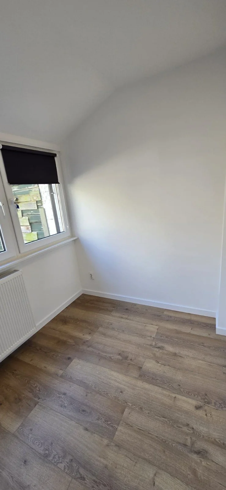 Leeg wit kamertje met houten vloer, twee ramen met zwarte rolgordijnen, radiator en witte muren.