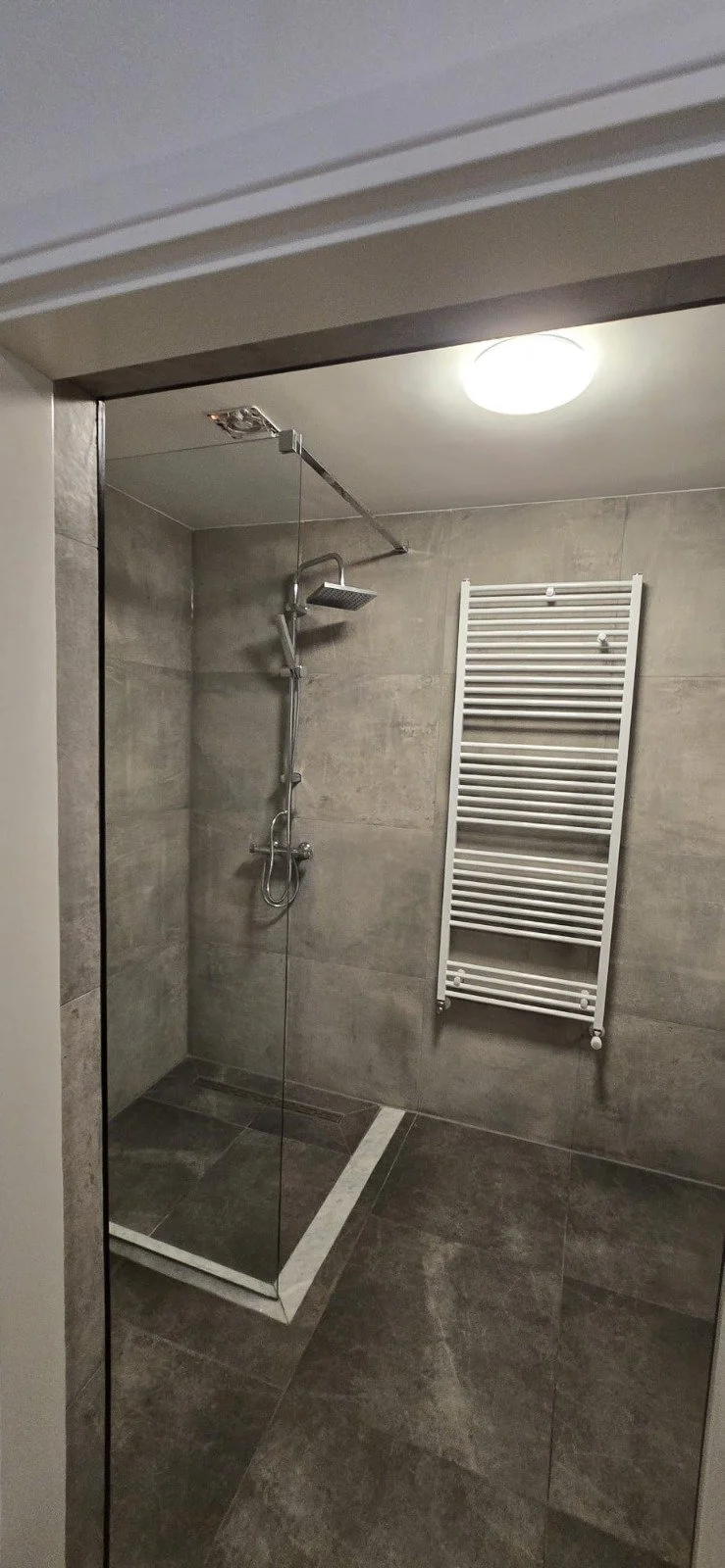 Moderne inloopdouche met bekleed met grote grijze tegels, een regendouchekop, een handdouche, en een witte radiator terras. Een plafondlamp verlicht de ruimte.