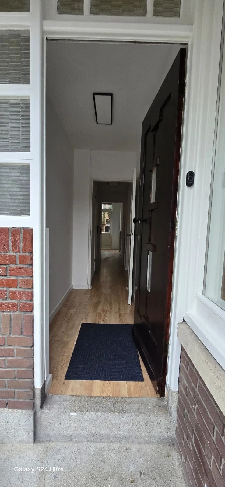 Een nieuwe woning met een openslaande voordeur, een donkere deur binnen, een houten vloer, een zwart matje en wit geschilderde muren.