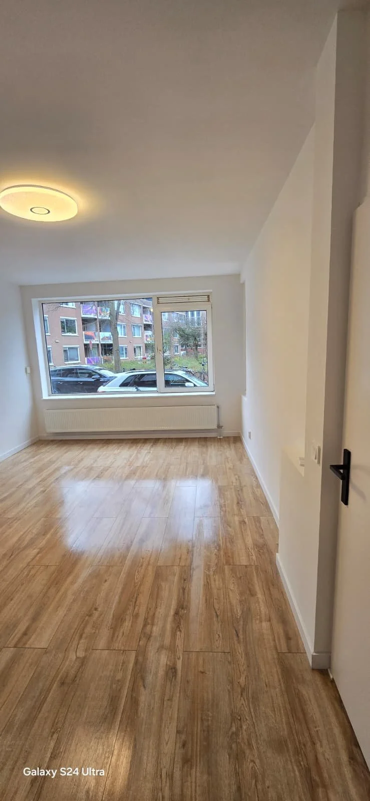 Lege kamer met grote raam, houten vloer, witte muren en plafondlamp, uitzicht op straat met auto's en appartementengebouw.