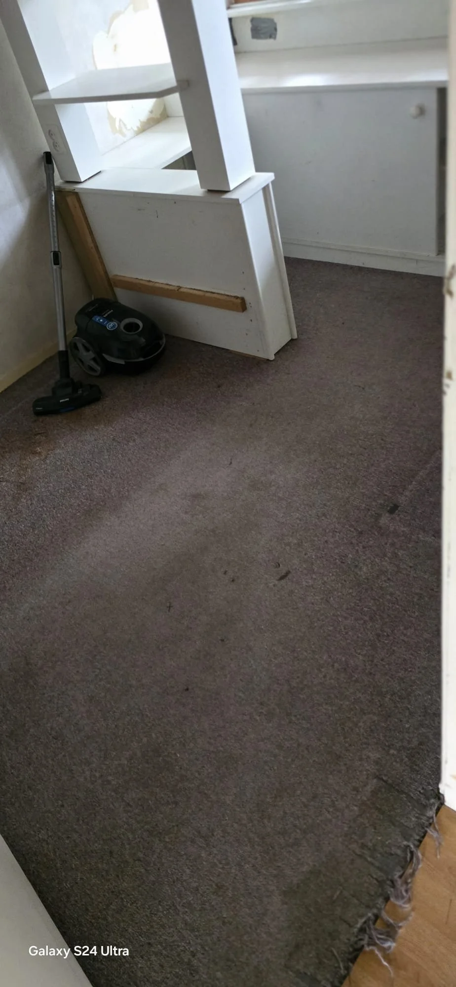 Een kamer met een stoffig tapijtvloer, een stofzuiger en een houten plank die op de vloer ligt. Er lijkt enige renovatie of schoonmaakwerkzaamheden te worden gedaan.