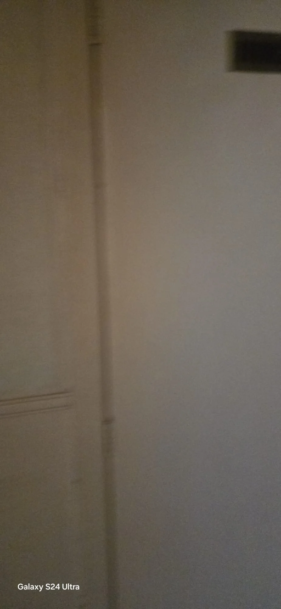 Close-up van een muur en deur met een donker vakje in de rechterbovenhoek, afbeelding gemaakt met een Galaxy S24 Ultra.