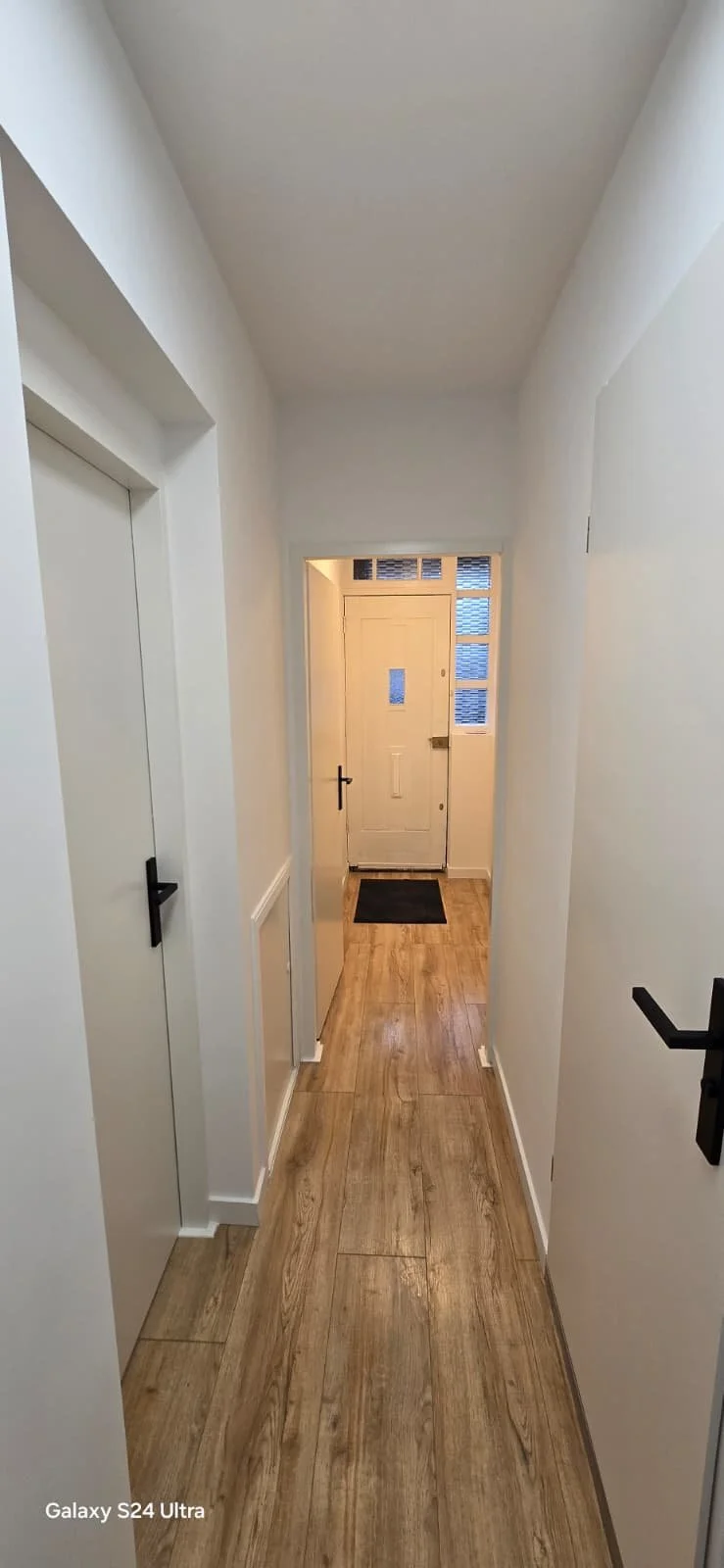 Een lange, smalle gang met houten vloer, witte muren en een witte voordeur aan het einde met ramen erboven. Links en rechts bevinden zich deuren.