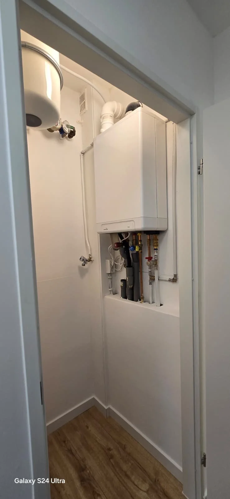 Een kleine kast met een boiler en wateraansluitingen in een witte kamer.