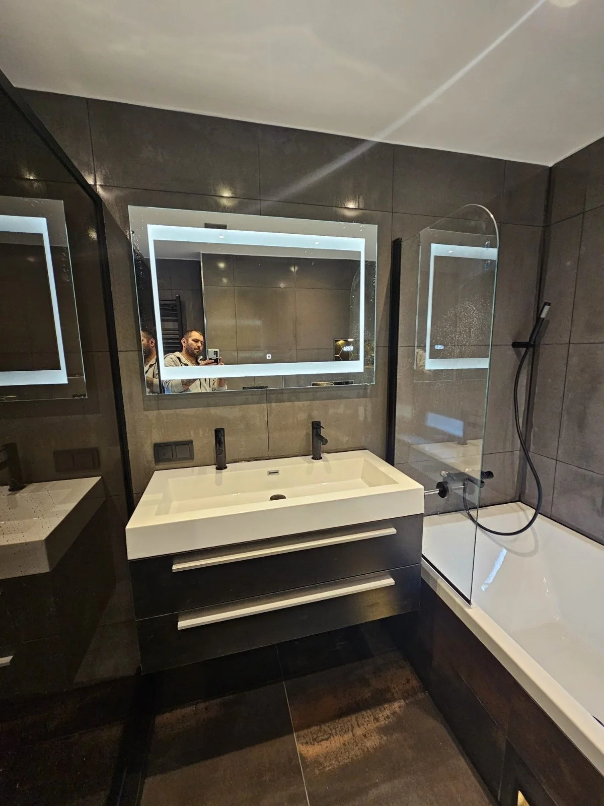 Een moderne badkamer met grote spiegel met verlichting, een wit wastafel met twee kranen, een bad met glazen douchewand, en donkere tegels op de muren en vloer.