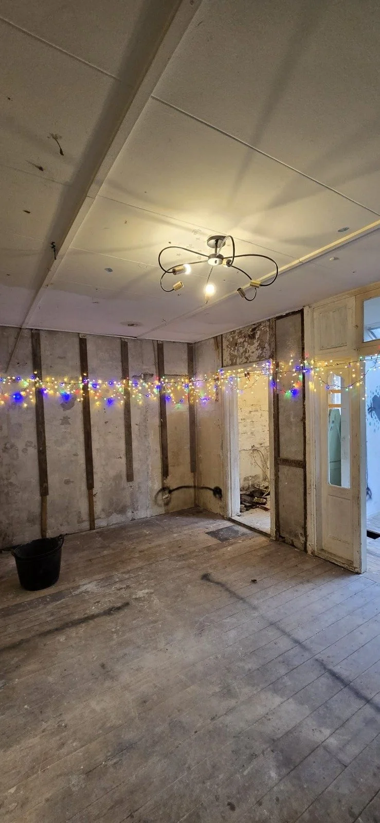 Een leeg, geruïneerd kamer met onafgewerkte muren, een versleten houten vloer, een plafondlamp met meerdere armaturen, en kerstversieringen met kleurrijke lichtjes langs de muur.