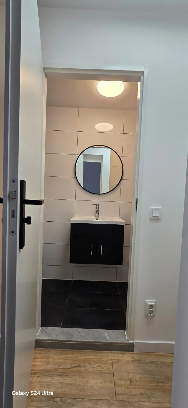 Een witte kamer met een open deur, binnenin een badkamer met een ronde spiegel boven een zwart badmeubel en een vaste wastafel, een plafondlamp, witte tegels op de achtergrond, en een stopcontact op de muur.