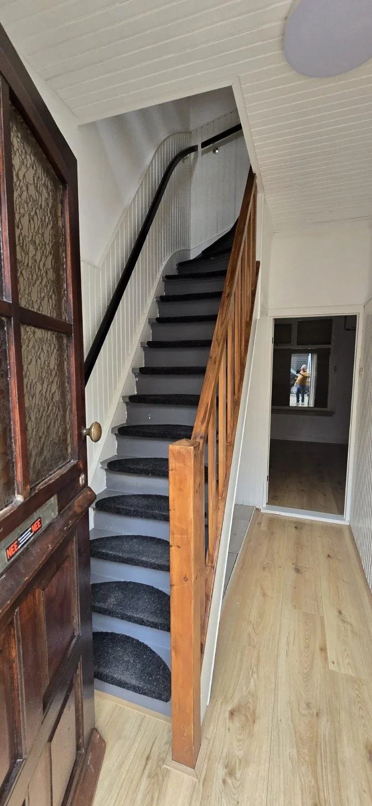 Binnenkant van een huis met een houten trap met zwart tapijt, witte wandpanelen, een raam met decoratief glas in de deuropening en een houten vloer.