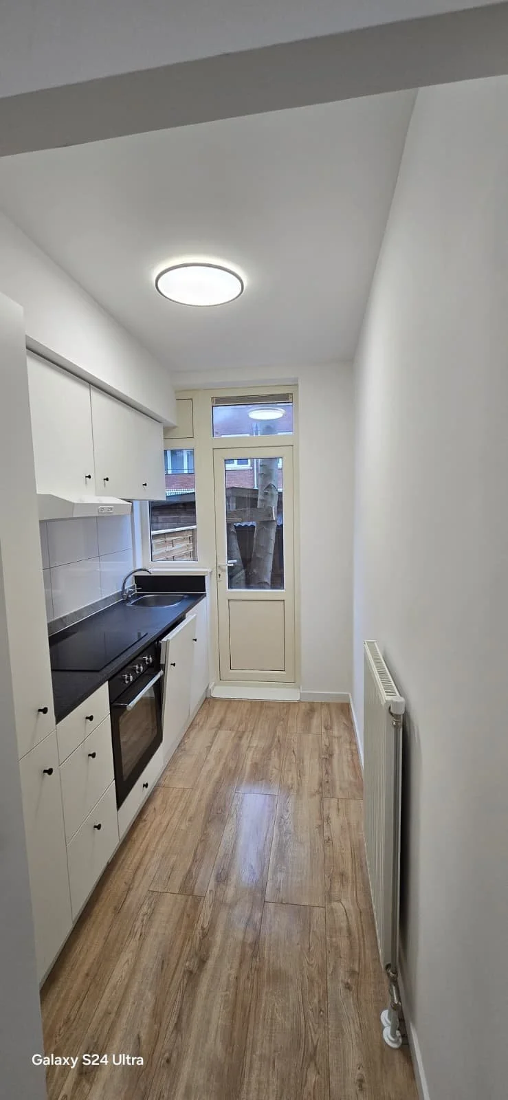 Kleine keuken met witte kasten, zwarte keukenblad, oven, gootsteen, deur naar buiten en radiateur aan de muur.