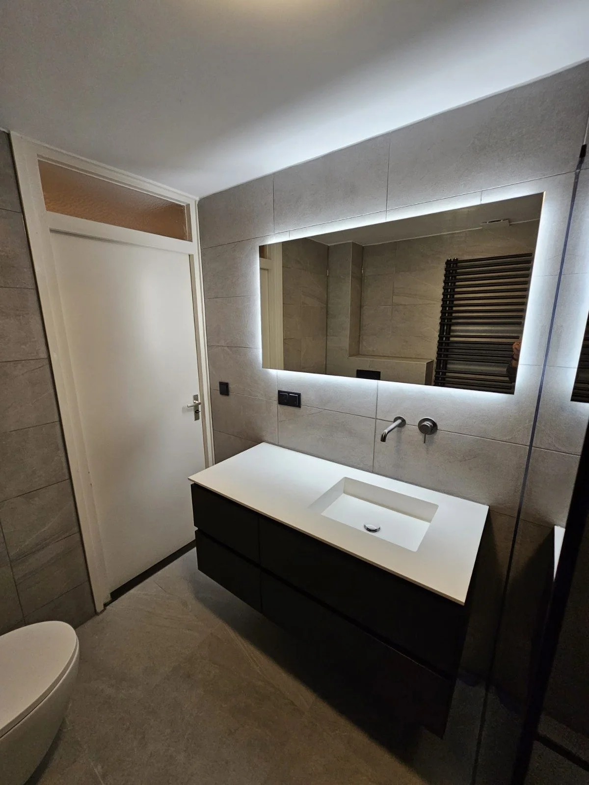 Moderne badkamer met een grote spiegel met verlichting, een witte wastafel met donker onderkast, een grote muur met beige tegels, en een toilet aan de linker zijde.