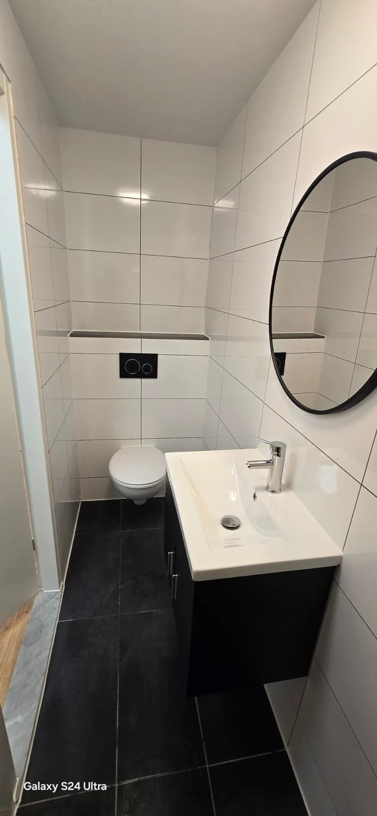 Een kleine, moderne badkamer met witte tegels en een zwart vloertje. Er is een wit wasbak met een spiegel ernaast en een muurontlasting.