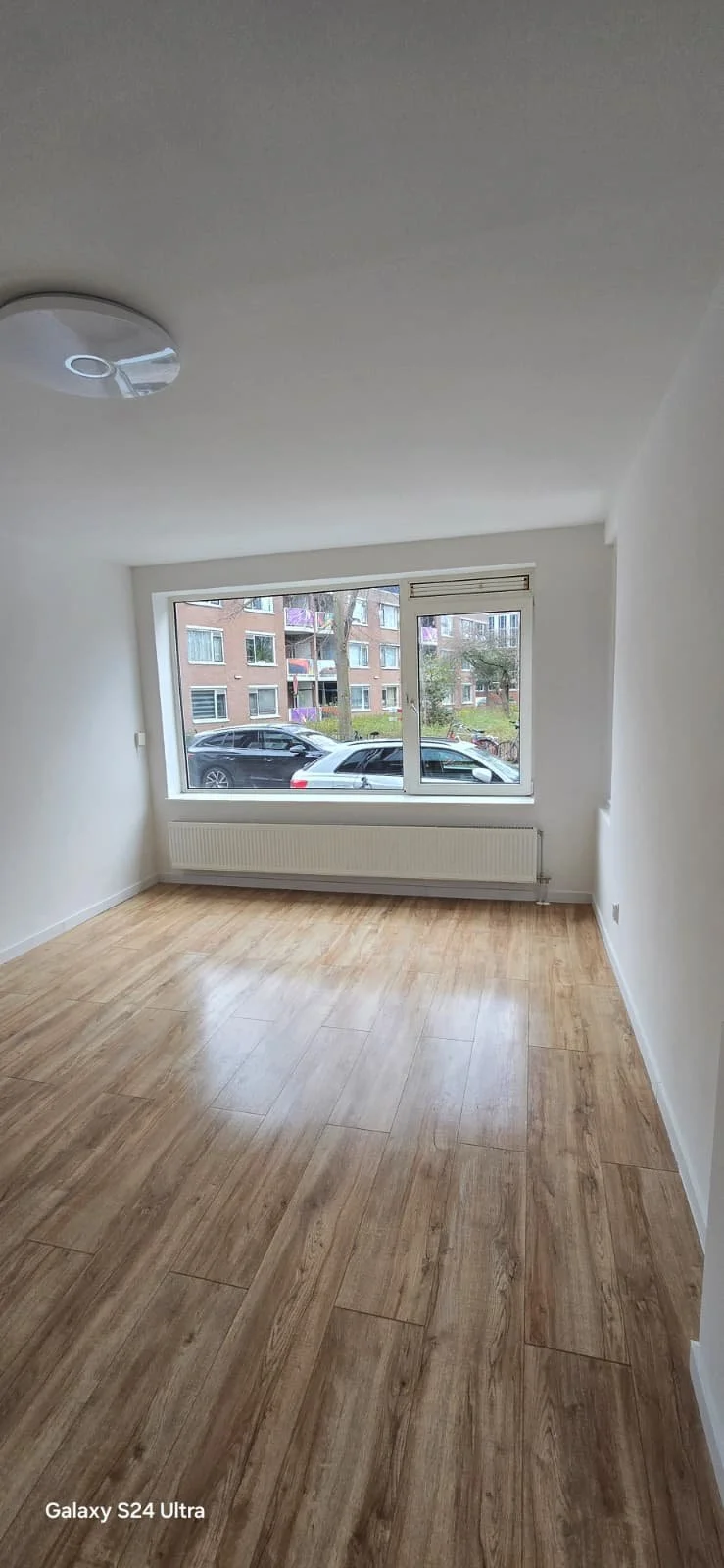 Leeg wit kamertje met houten vloer, grote raam en radiator.