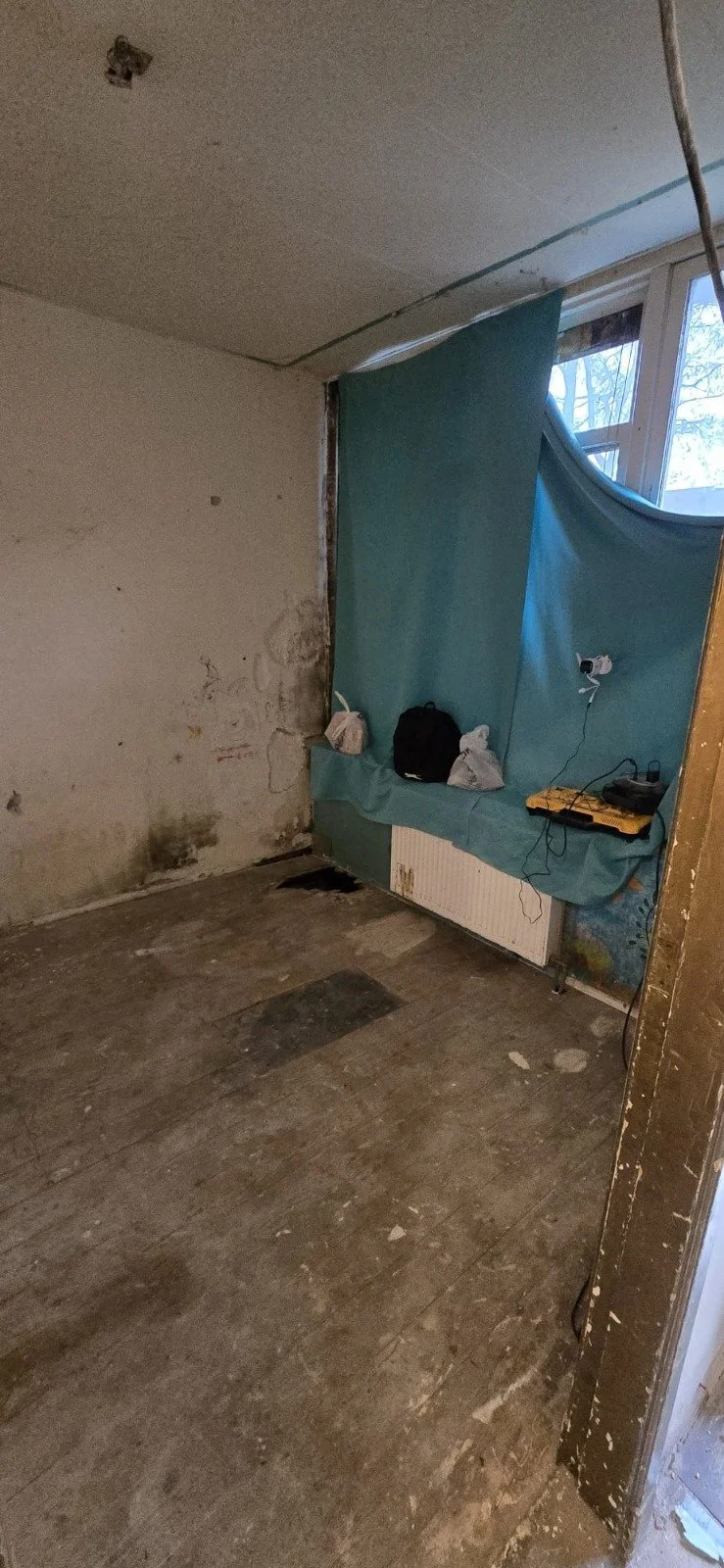 Een kamer die gerenoveerd wordt met onafgewerkte muren en vloer, een raam met een blauwe gordijn, een radiator, en enkele spullen op een tafel.