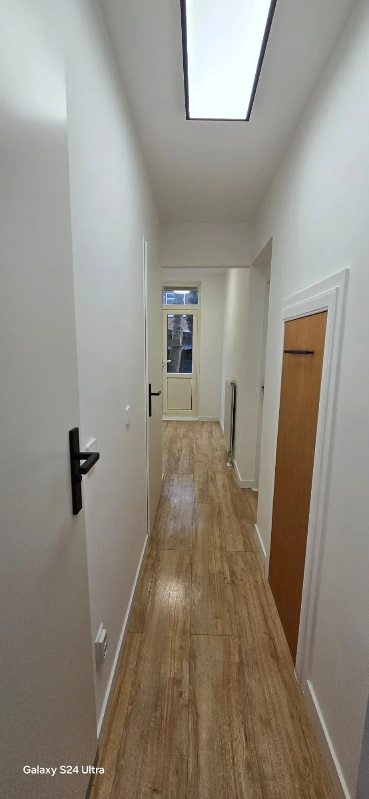 Lange, smalle gang met houten vloer, witte muren, een plafondlicht, een raam en een deur aan het einde.