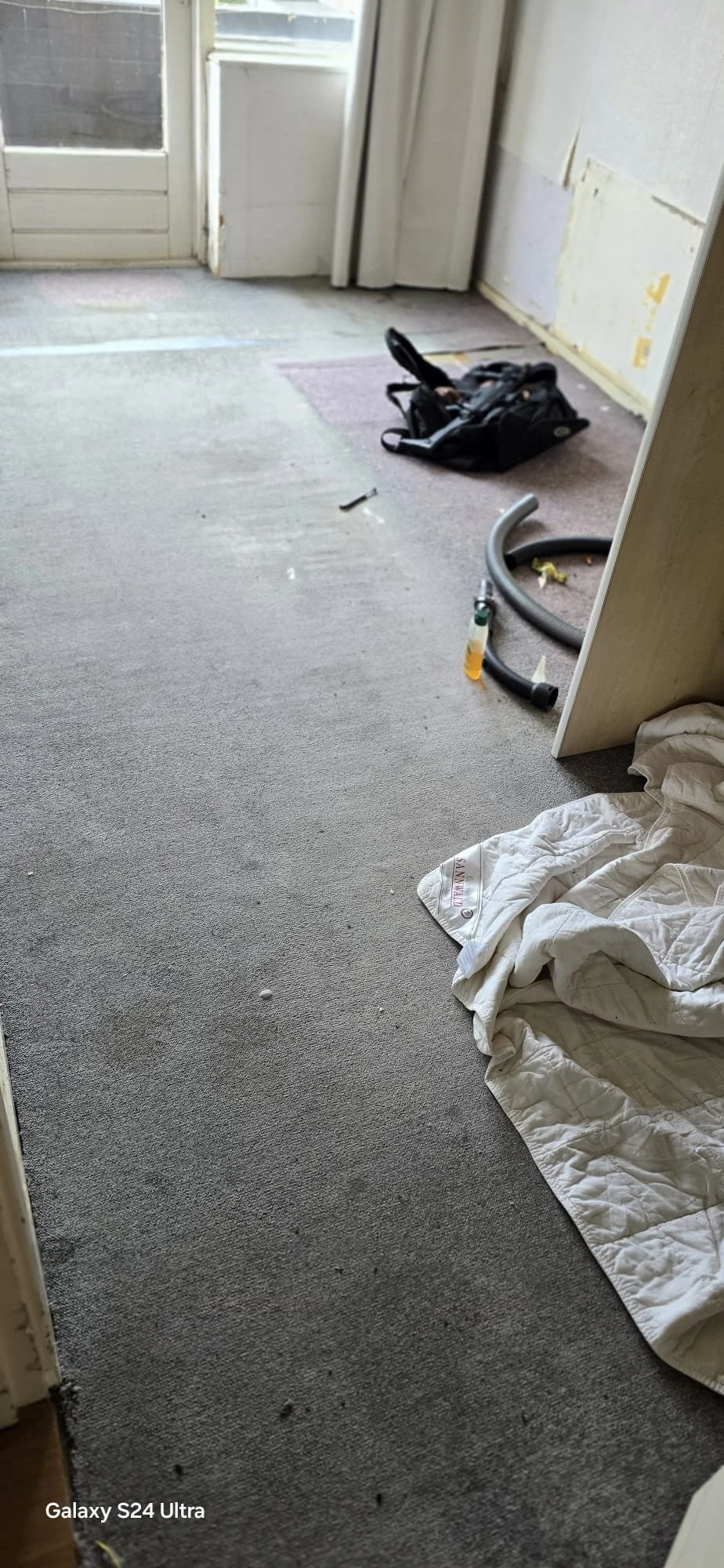 Een kamer onder renovatie met een open deur, een vloerbedekking, een zwart rugzak, een stuk plastic, een stofzuigerslang, een doek op de vloer en een bedlok.