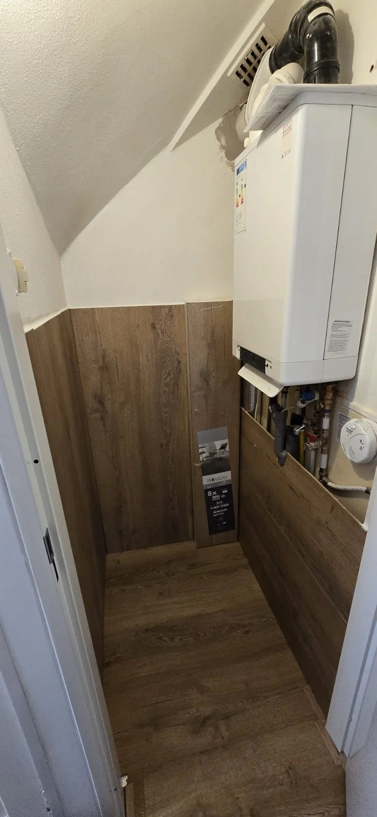 Een kleine wasruimte met een wasmachine en een verwarmingsketel, afgewerkt met houten wandpanelen en een houten vloer.