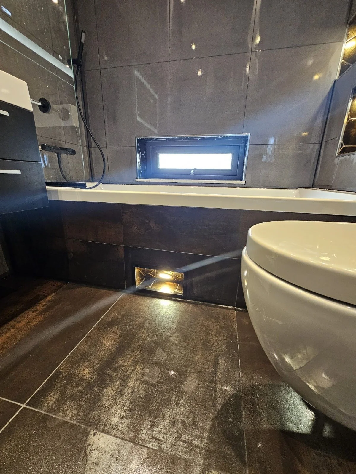 Moderne badkamer met donkere tegels, een wit toilet, een klein raam, en ingebouwde verlichting.