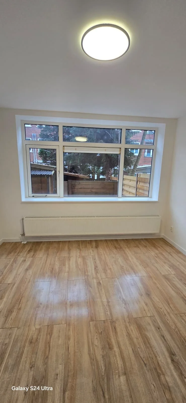 Leeg kamertje met houten vloer, grote mannelijke raam, witte muren en plafond met ingebouwde lamp.