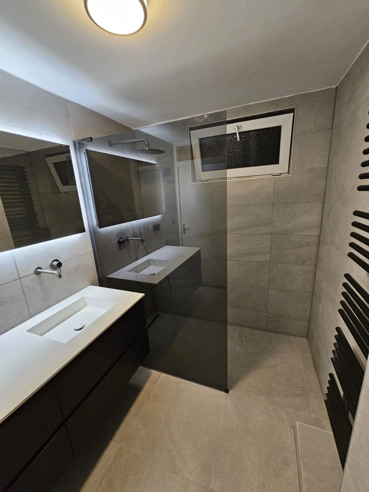 Moderne badkamer met dubbele wastafel, grote spiegel, inloopdouche met glazen wand, ventilatierooster in raam, centrale plafondlamp en verwarmingsradiator.