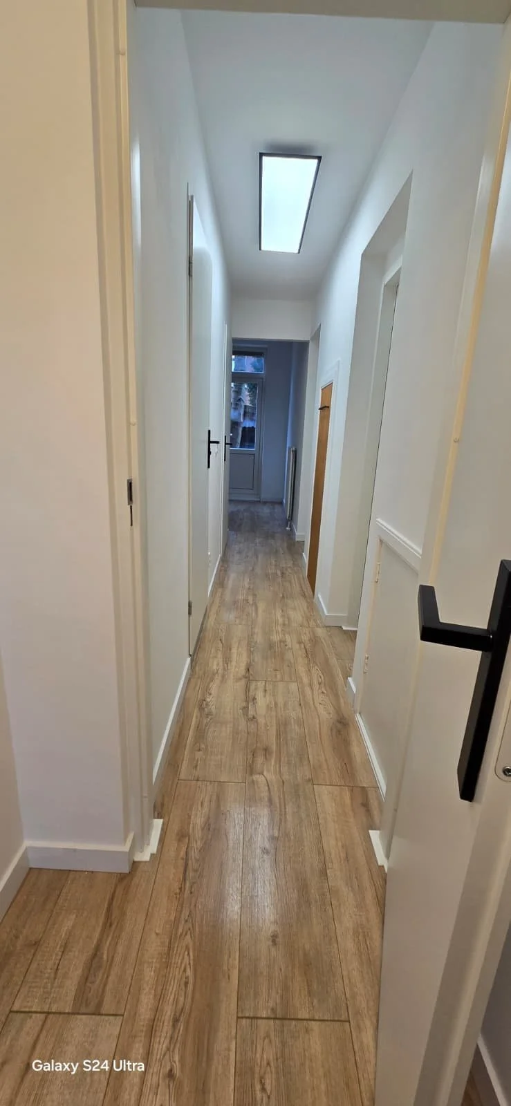 Binnenkijkend in een smalle witte gang met houten vloer, plafond met ingebouwd licht en een raam aan het einde.