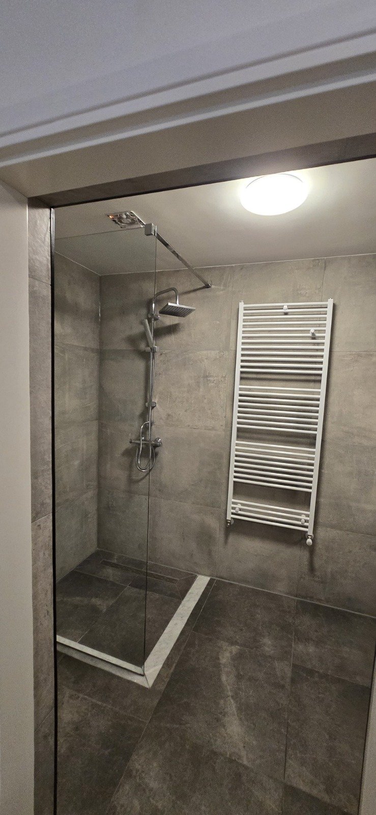 Badkamer met een inloopdouche, stortdouche met plafondpulsensor, witte verwarmingsradiator aan de muur, plafondlamp, betegelde muren en vloer.