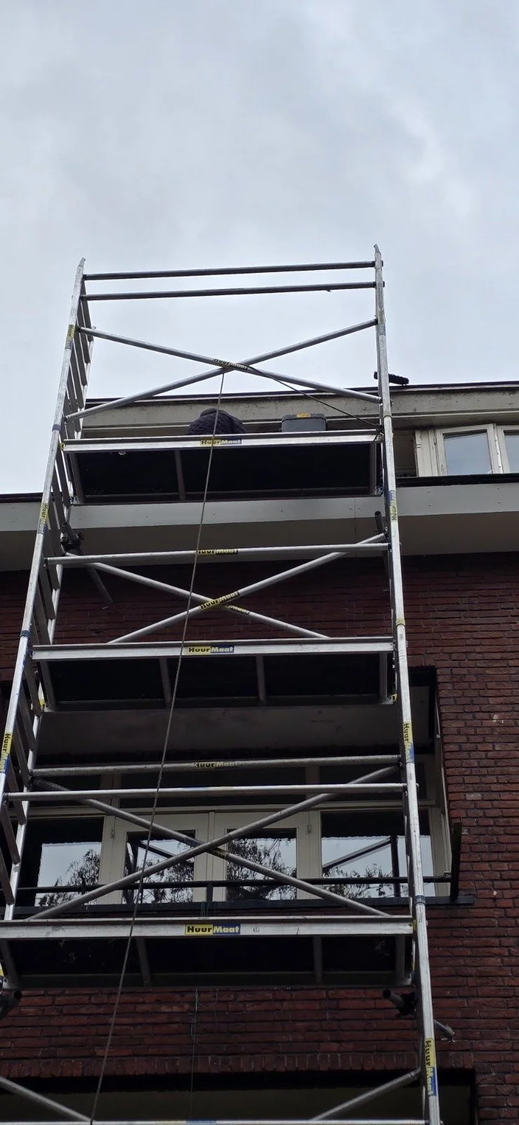 Stalen steiger voor gevelrenovatie voor een gebouw met een rood bakstenen gevel en enkele ramen, onder een overhang en onder een grijze lucht.