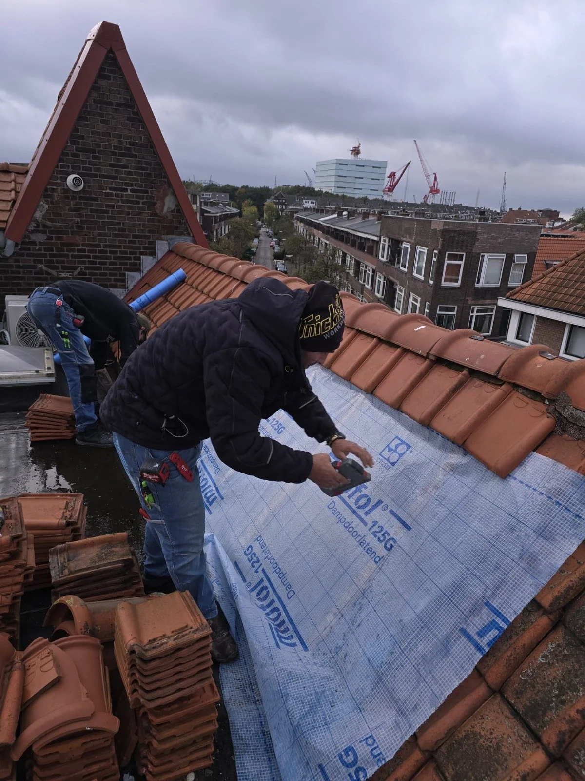 Twee bouwvakkers werken op een schuine daken met dakpannen, een van hen legt isolatiemateriaal aan.