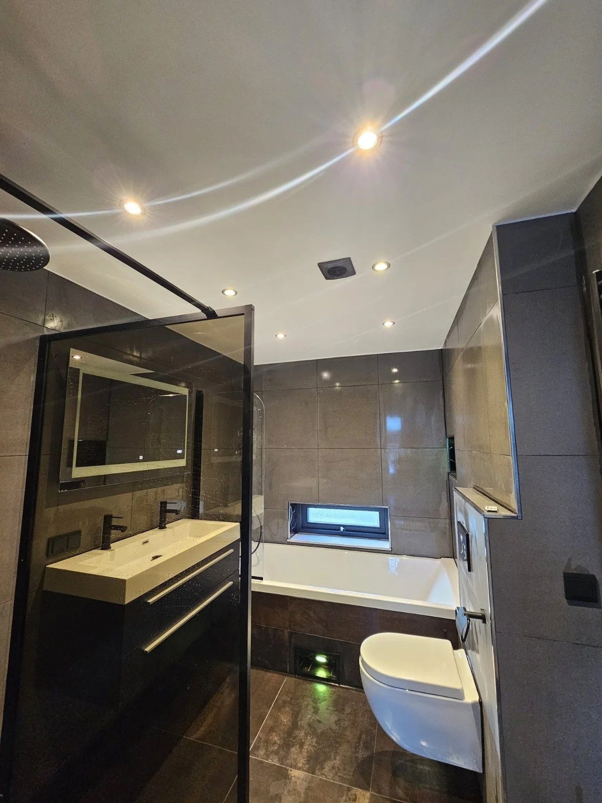 Moderne badkamer met dubbele wastafel, spiegel, inloopdouche, bad en toilet, met donkere betegeling en ingebouwde verlichting.