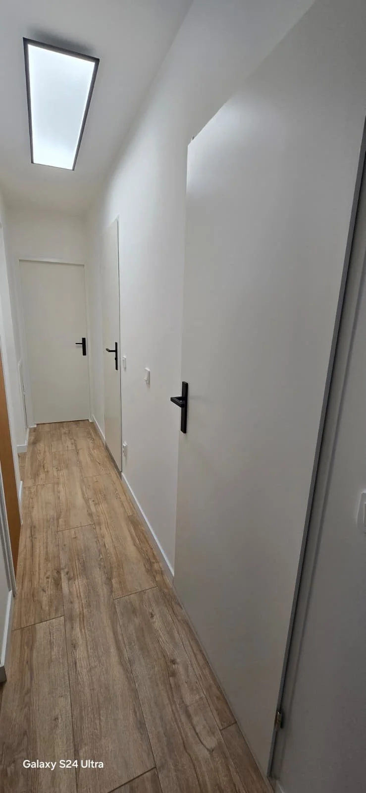 Kleine gang met witte muren, houten vloer, witte deuren en zwarte deurknoppen, verlicht door een rechthoekig plafondlicht.