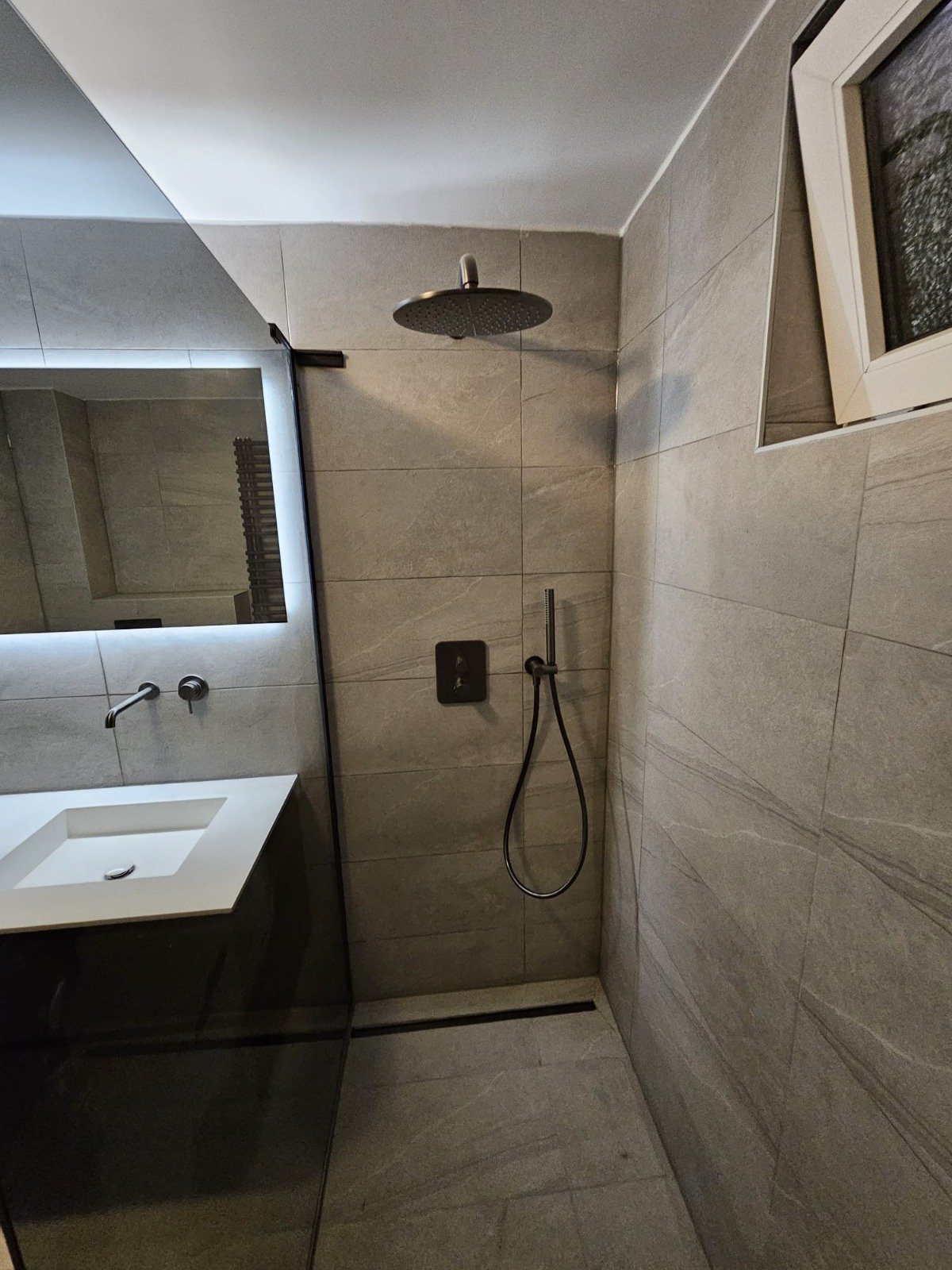 Moderne badkamer met inloopdouche, grote grijze tegels, wastafel met spiegel en wandhouder in zwart.