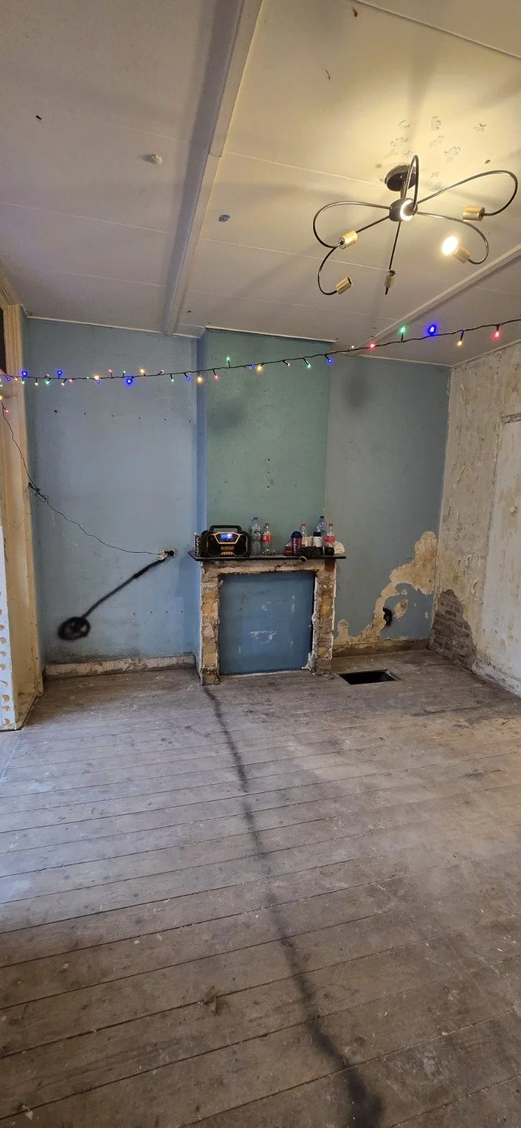 Leeg kamertje met een beschadigde muur, een oude houten vloer, en een plafond met een moderne kroonluchter en kerstverlichting. Er staan flesjes op een schoorsteenmantel.